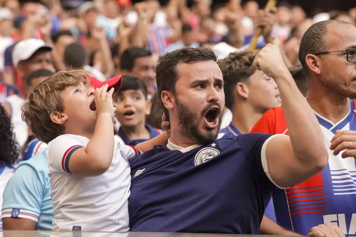 A football stadium with spectators in the stands, some silhouettes with arms raised, evening light filtering through the stadium structure, crowd energy captured in motion blur.