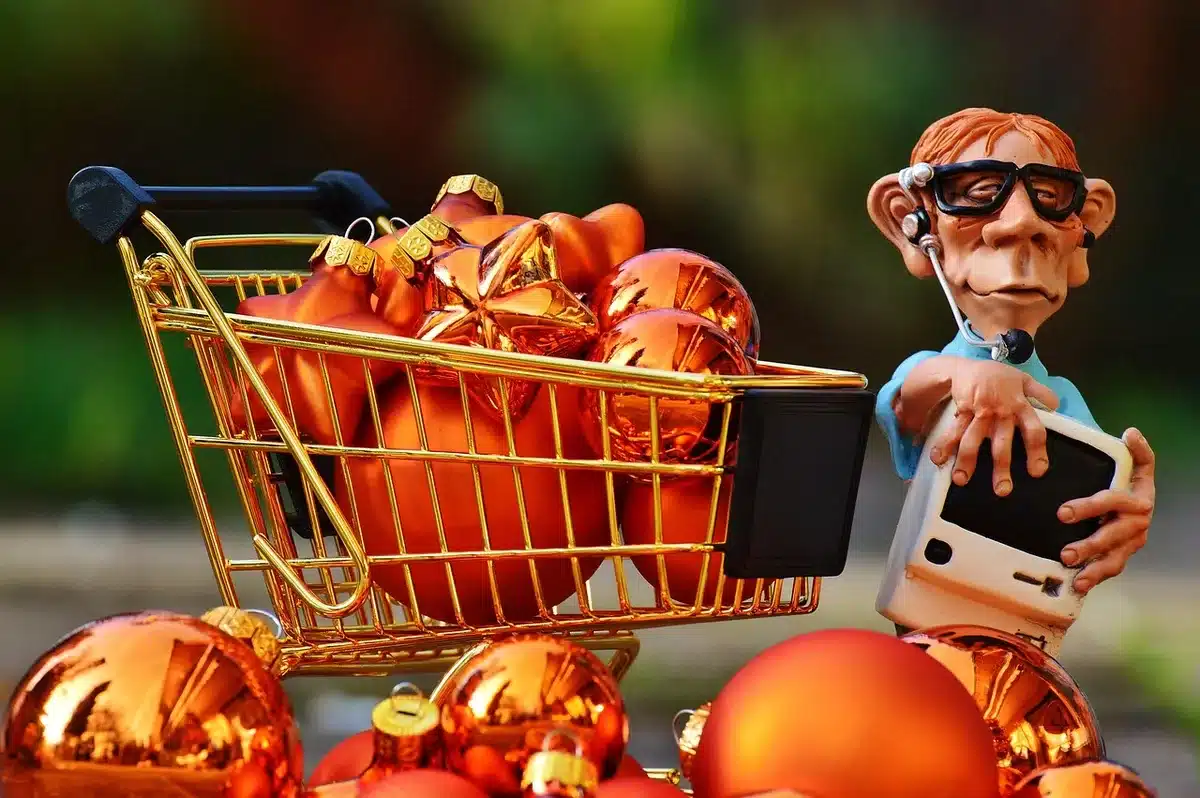 Close-up of an anonymous hand holding a smartphone over a laptop showing a shopping cart page, blurred product listings in the background