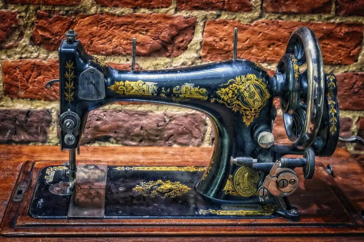 Close-up of a mechanical sewing machine with white and pink details, showing the stitch selector dial, thread spool, and presser foot on a clean studio surface, surrounded by fabric scraps and thread colors